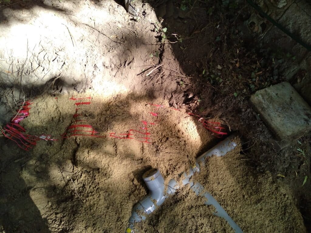 Tranchée de sable avec des tuyaux de raccordement pour un réseau souterrain en cours d'installation, entourée de végétation et de terre.