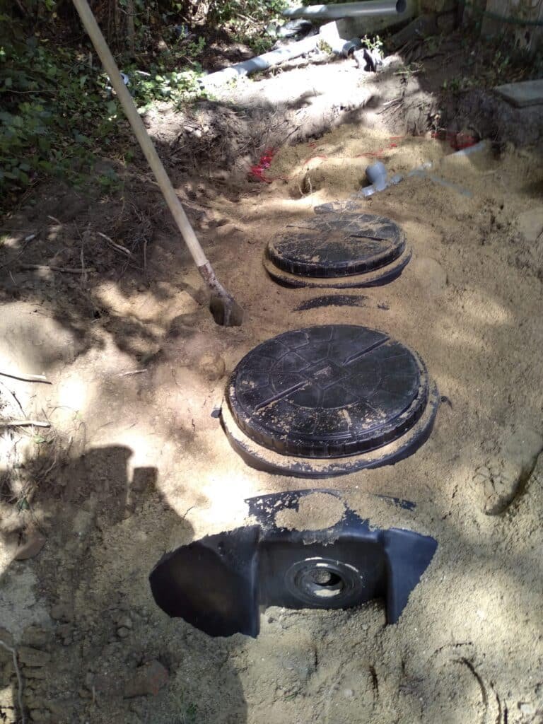 Vue de deux couvercles noirs en plastique installés dans une tranchée de sable, dans le cadre d'un projet d'installation d'un système de canalisation souterrain.