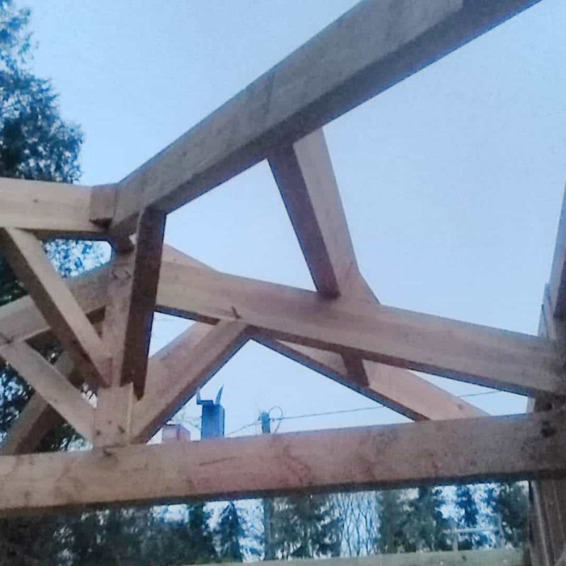 Vue rapprochée d'une charpente en bois d'une maison en construction, avec des poutres croisées et un ciel légèrement nuageux en arrière-plan.