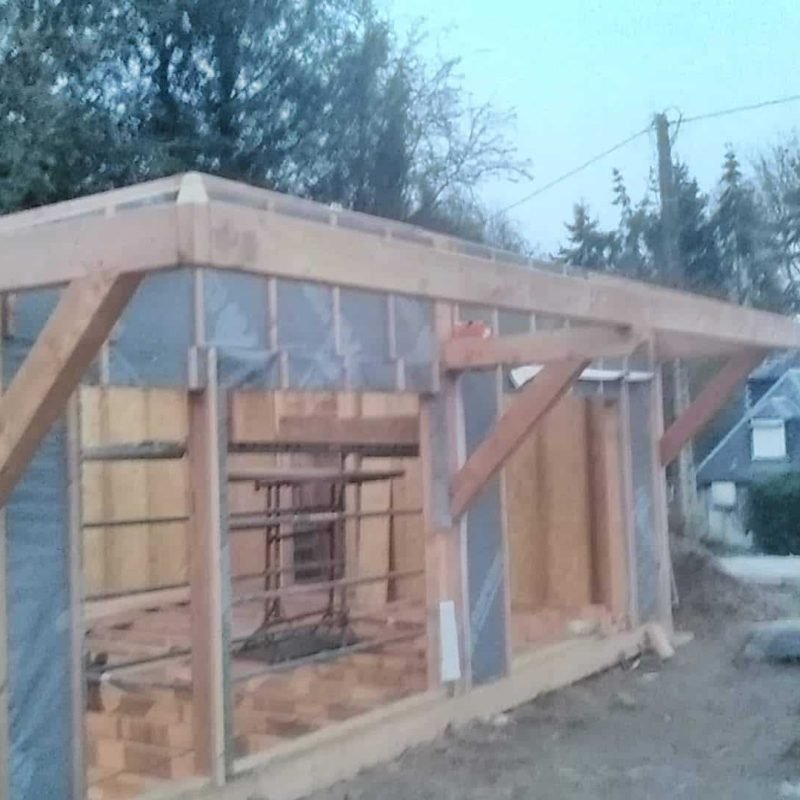 Structure en bois d'une maison en cours de construction avec des échafaudages et des poutres visibles, en arrière-plan d'une zone résidentielle.
