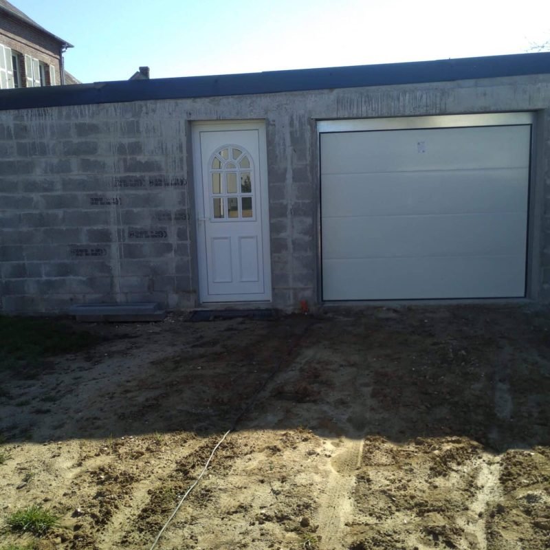 Garage en construction avec des murs en parpaings, une porte d'entrée blanche à gauche et une porte de garage blanche à droite, sur un terrain non pavé.