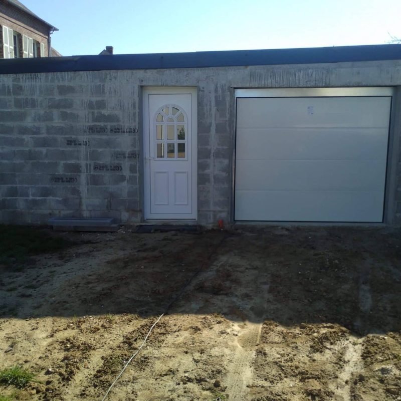 Garage en construction avec des murs en parpaings, une porte d'entrée blanche à gauche et une porte de garage blanche à droite, sur un terrain non pavé.
