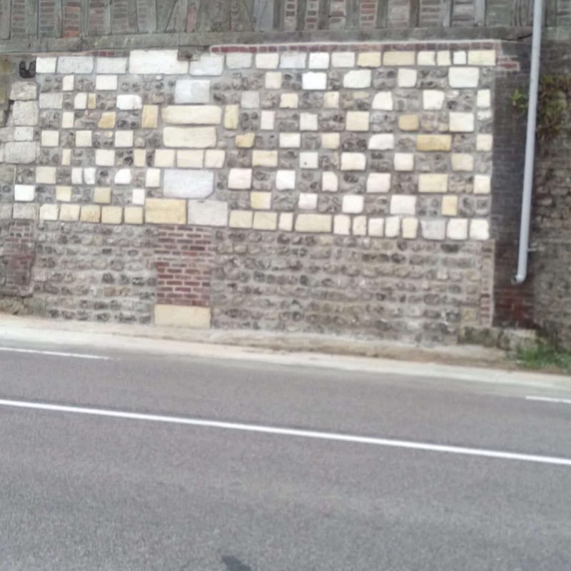 Vue d'un mur en pierre et briques rénové avec un motif géométrique de différentes tailles et couleurs, longeant une route.