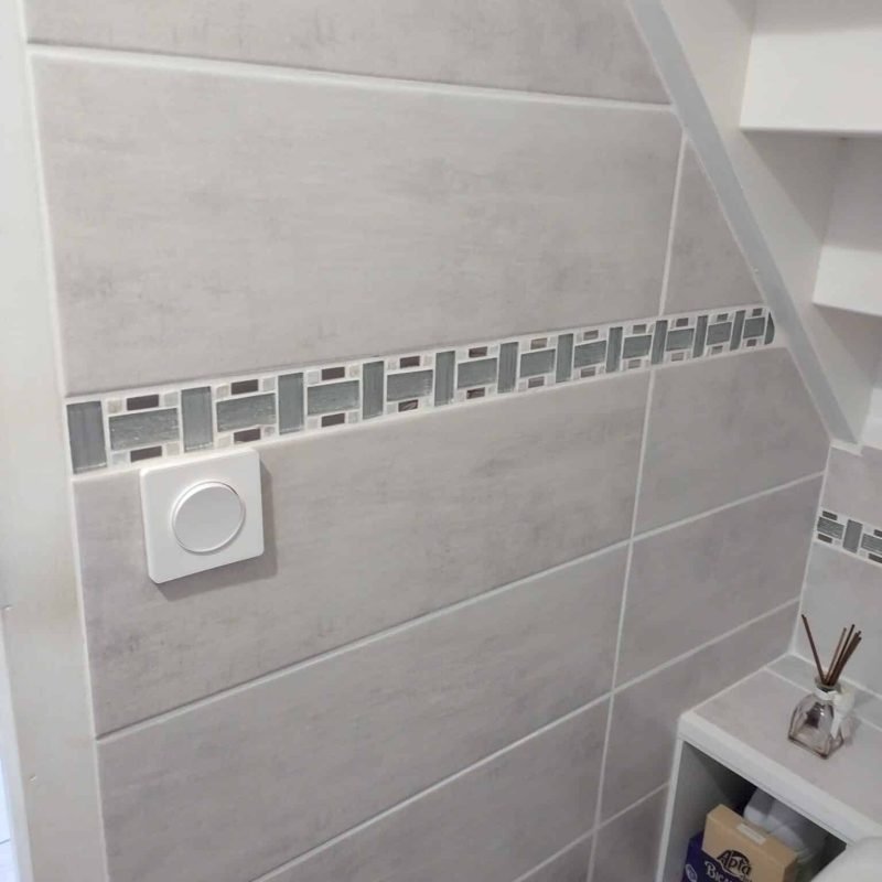 Détail d'un mur carrelé dans une salle de bain, avec un interrupteur et des carreaux décoratifs.