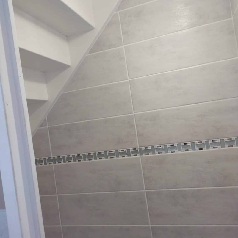 Vue d'un mur de salle de bain carrelé avec une bande décorative sous un escalier.