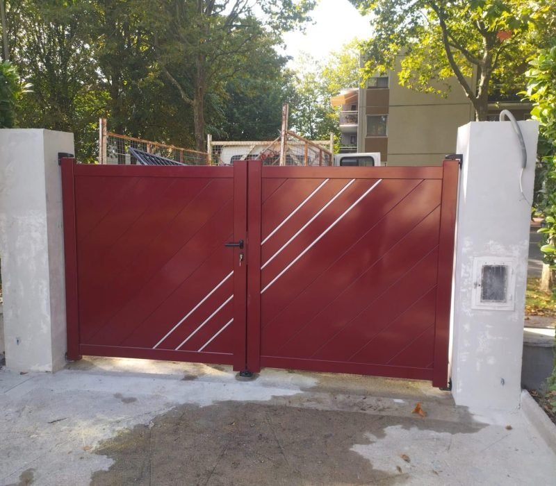 Nouveau portail moderne en aluminium installé dans une entrée résidentielle Portail en aluminium de couleur rouge avec des lignes diagonales en métal, installé entre deux piliers en béton dans une entrée résidentielle, entouré de verdure