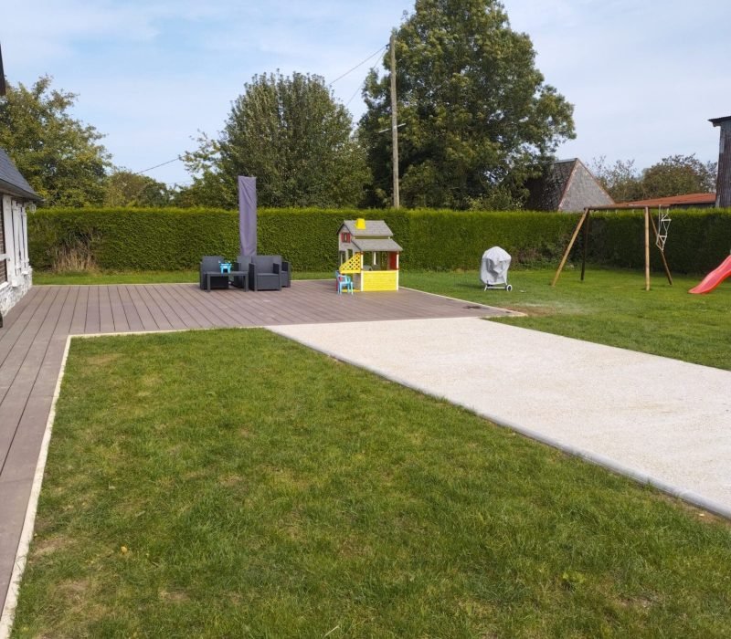 Aménagement d’un jardin avec terrasse en bois et espace de jeu pour enfants Jardin résidentiel bien entretenu avec une grande terrasse en bois, une pelouse et une aire de jeu pour enfants comprenant une maison de jeu et une balançoire, entouré de verdure.