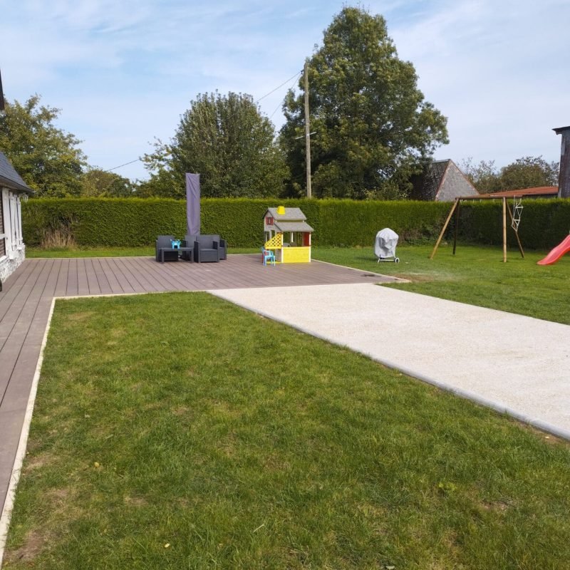 Jardin résidentiel bien entretenu avec une grande terrasse en bois, une pelouse et une aire de jeu pour enfants comprenant une maison de jeu et une balançoire, entouré de verdure.