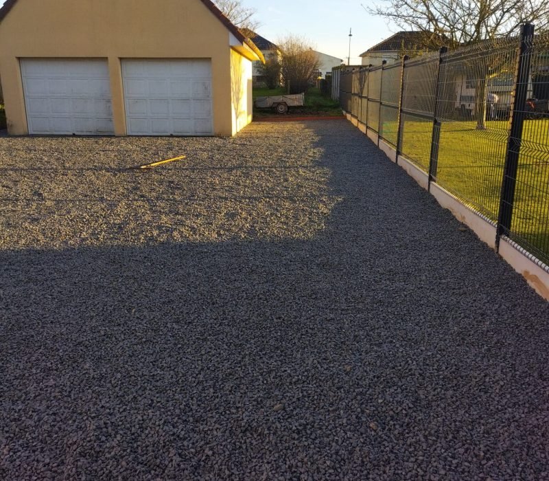 Aménagement d’une cour en gravier devant un garage résidentiel Vue d'une cour en gravier récemment installée devant un garage double, avec une clôture métallique sur le côté droit, dans un cadre résidentiel ensoleillé.