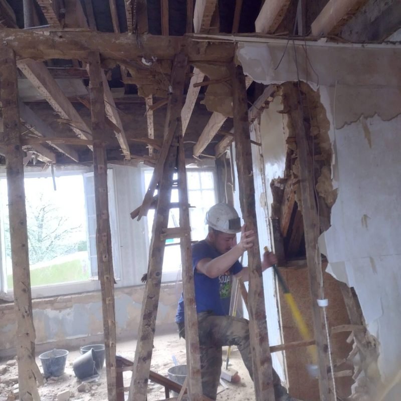 Un ouvrier portant un casque de sécurité effectue des travaux de démolition à l'intérieur d'un bâtiment, retirant des éléments de structure et de plâtre, entouré de poutres en bois.