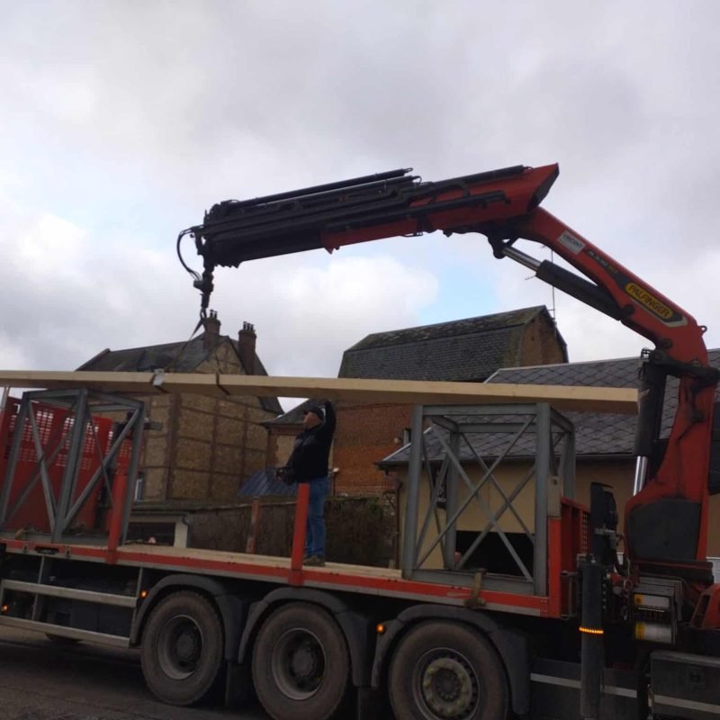 Un ouvrier guidant la grue d'un camion pour soulever et positionner une poutre en bois destinée à un chantier de construction ou de rénovation, avec un camion équipé d'une grue hydraulique
