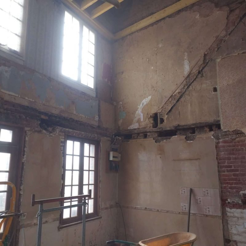Travaux de rénovation intérieure montrant une pièce avec des murs abîmés, des fenêtres en bois et un échafaudage, dans le cadre d'une réhabilitation structurelle.